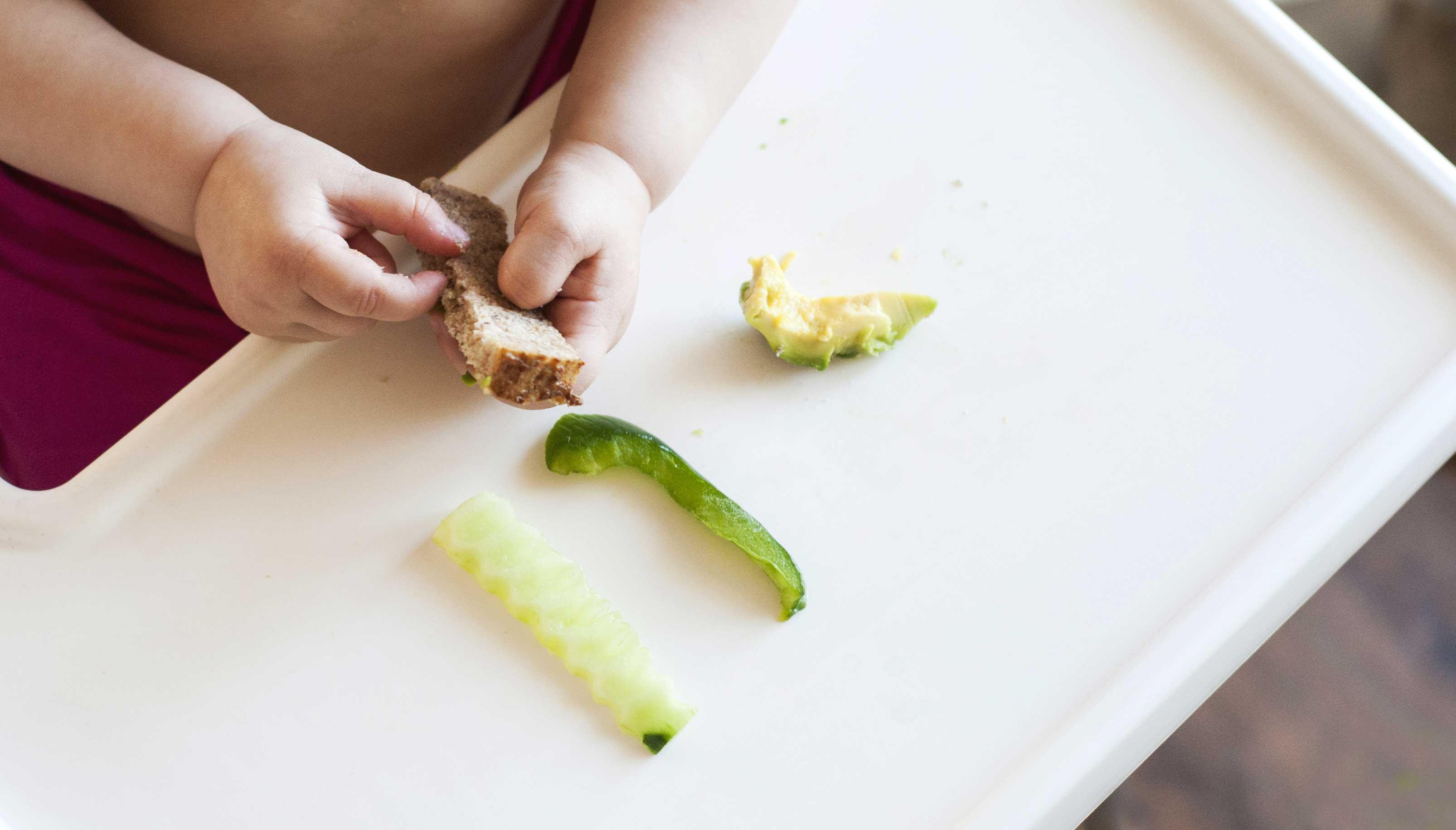 Baby hält ein Brotstäbchen in den Händen. Auf dem Tisch liegen 3 grüne Gemüse in Streifen geschnitten